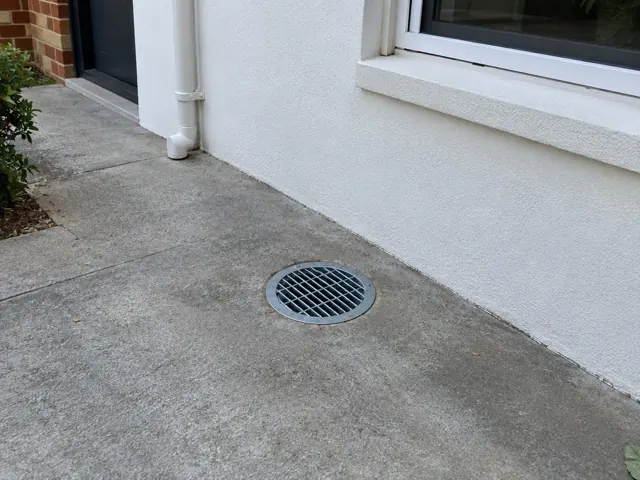 Outdoor concrete path with a round drain grate beside a white house wall and window in Killara