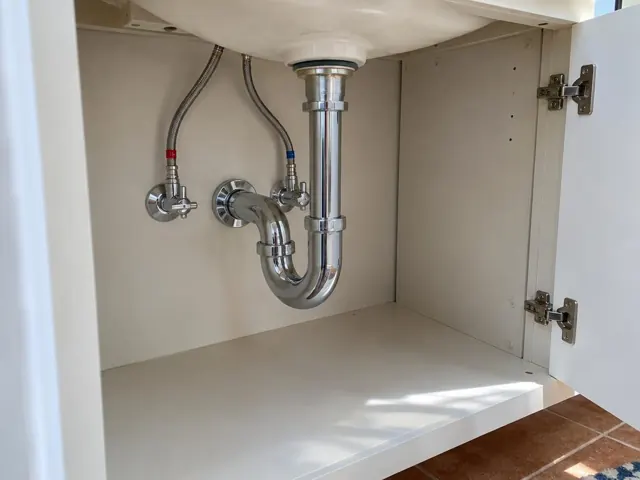 Open under-sink cabinet with polished drain pipe, shutoff valves and white access panel in Lindfield